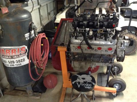1985 Jeep CJ7 - A V8 engine is mounted on an orange engine stand, with an air compressor and a red air hose to the left. A black rolling stool with a skull…