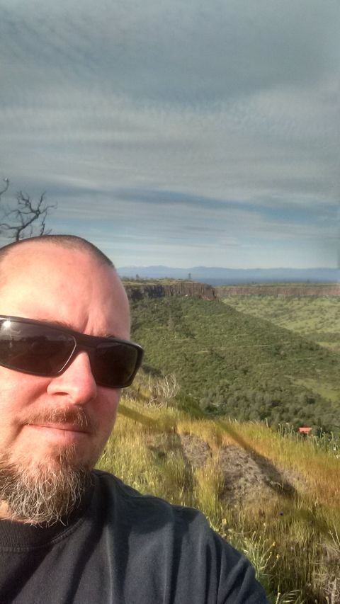1985 Jeep CJ7 - A man takes a selfie in front of a scenic overlook with a canyon and rolling green hills under a cloudy sky.