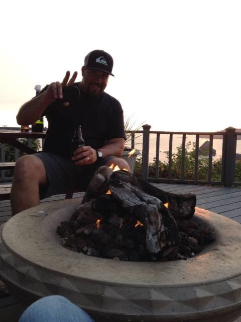 1985 Jeep CJ7 - A man wearing a black baseball cap and t-shirt holds up a smartphone and a beer bottle while sitting by a fire pit on a deck. The backgroun…