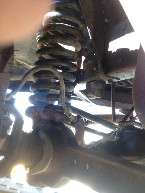 1979 Ford Bronco - The underside of a vehicle shows a heavily rusted suspension system, including a large coil spring and various metal components. A brake li…