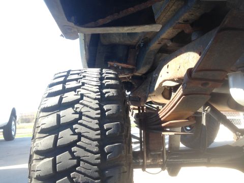 1979 Ford Bronco - A low-angle shot of the undercarriage of a vehicle, focusing on the large, knobby tire and rusty leaf spring suspension system. A portion o…