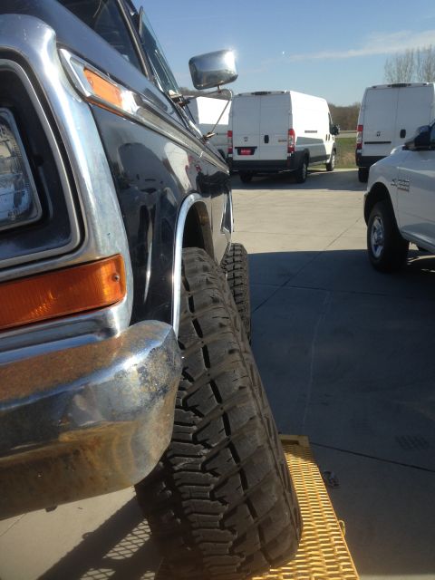 1979 Ford Bronco - Close-up, low-angle side view of the front fender and large tire of a dark-colored vehicle. In the background, several white cargo vans are…