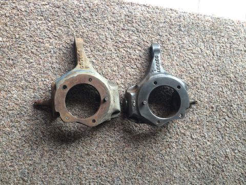 1981 Ford Bronco - Two steering knuckles are shown side-by-side on a carpeted surface. The knuckle on the left appears older and shows some rust and blue pain…
