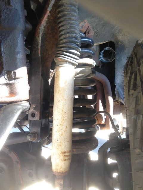 1979 Ford Bronco - A close-up view of the front suspension components of a vehicle, featuring a coil spring, shock absorber, and various rusted metal brackets…