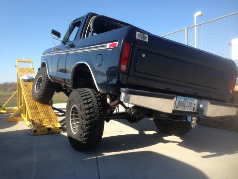 1979 Ford Bronco - The rear driver's side of a lifted, dark blue 1979 Ford Bronco is shown from a low rear-three-quarter angle. The vehicle is on a yellow ram…