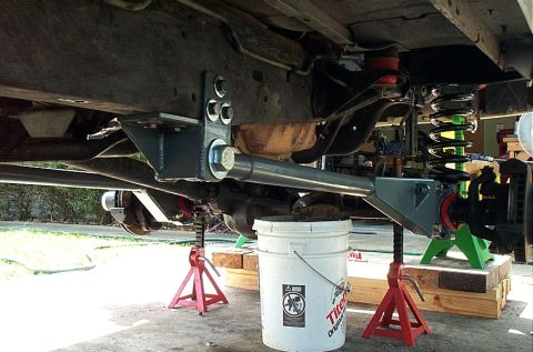 1995 Ford Bronco - Underside view of a vehicle raised on red jack stands and wood blocks, showing front suspension with a coil spring, control arm, and a larg…