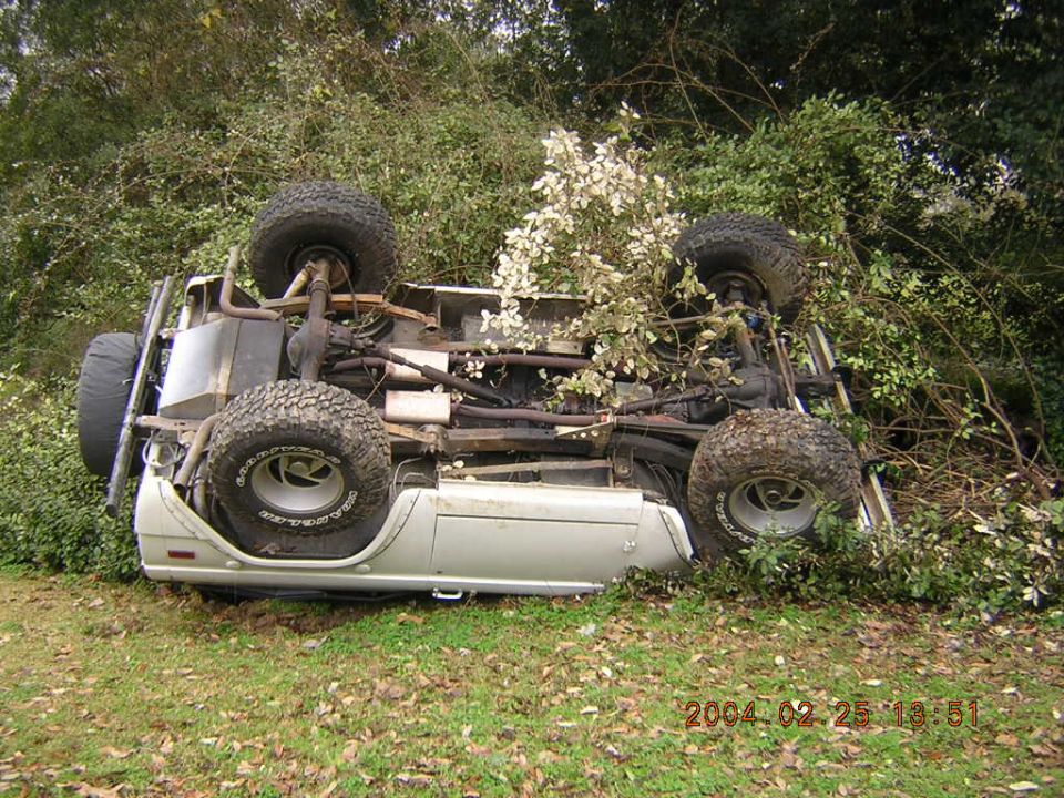 White SUV on its roof, partially obscured by foliage, with visible undercarriage and large tires