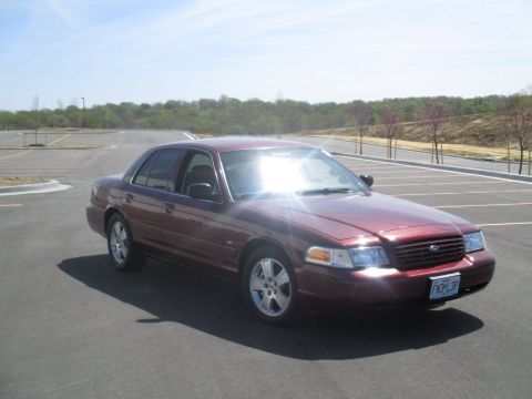2004 Ford Crown Victoria - A maroon 2004 Ford Crown Victoria sedan is parked in an empty asphalt parking lot under a clear blue sky. The car is viewed from a 3/4 fron…