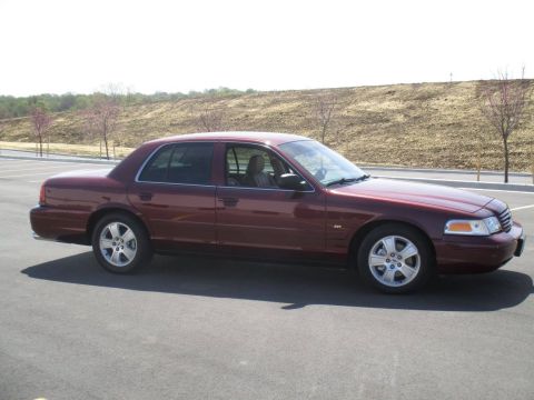 2004 Ford Crown Victoria - A maroon 2004 Ford Crown Victoria sedan is parked on an asphalt surface with a dirt embankment and sparse trees in the background.
