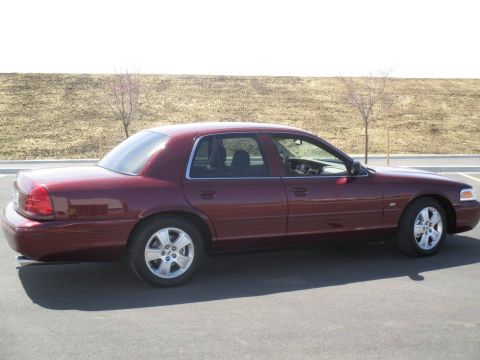 2004 Ford Crown Victoria - The side view of a maroon 2004 Ford Crown Victoria is shown parked on asphalt with a dirt embankment and sparse trees in the background.