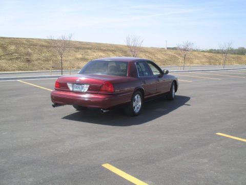 2004 Ford Crown Victoria - The rear passenger side of a dark red Ford Crown Victoria is parked in an empty asphalt parking lot. The car has a license plate that reads…