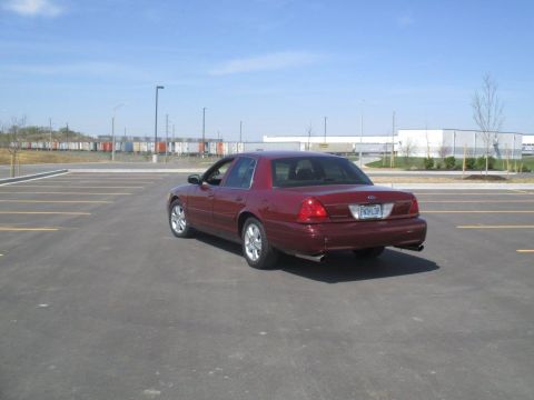 2004 Ford Crown Victoria - The rear passenger side of a dark red Ford Crown Victoria is shown parked in an empty asphalt parking lot. The car has a blue license plate…