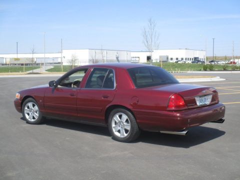 2004 Ford Crown Victoria - The rear passenger side of a dark red Ford Crown Victoria sedan is shown parked in an asphalt lot with buildings in the background. The veh…