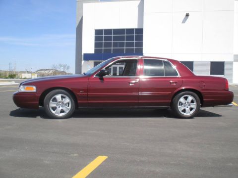 2004 Ford Crown Victoria - Side profile of a maroon Ford Crown Victoria sedan parked in front of a modern building with large windows. The car features chrome trim ar…