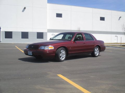 2004 Ford Crown Victoria - A maroon Ford Crown Victoria sedan is parked in a paved lot in front of a modern white building with dark windows. The car is viewed from a…