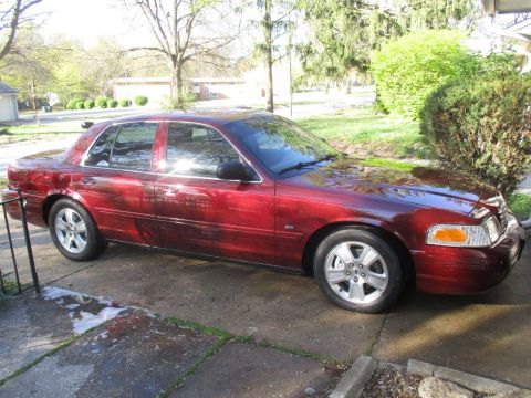 2004 Ford Crown Victoria - A shiny, dark red 2004 Ford Crown Victoria sedan is parked on a concrete driveway. The car is viewed from the side, showing its full length…