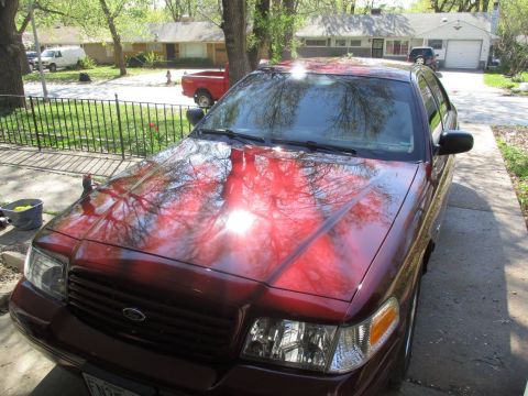 2004 Ford Crown Victoria - The front driver's side quarter view of a dark red Ford Crown Victoria is shown parked in a driveway. The car's hood and windshield reflect…