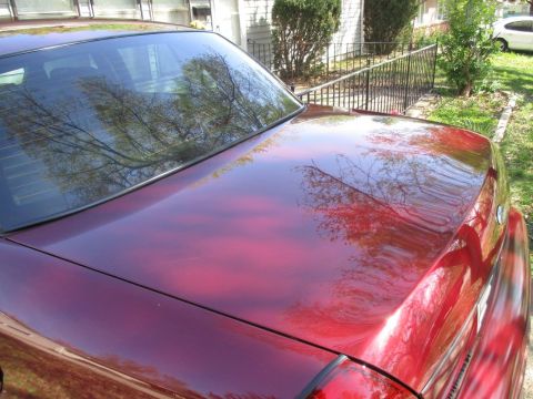 2004 Ford Crown Victoria - The rear passenger side of a dark red sedan is shown with reflections of trees and sky on its polished trunk and rear window. The car is pa…
