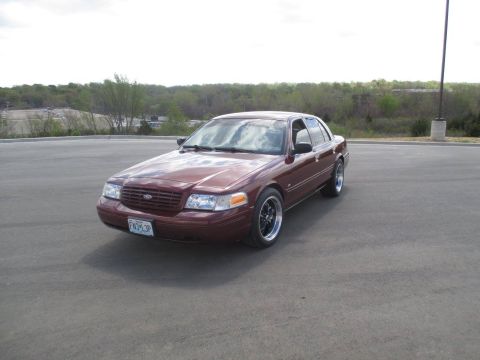2004 Ford Crown Victoria - A maroon 2004 Ford Crown Victoria sedan is parked on asphalt with a wooded area and cloudy sky in the background. The car has aftermarket b…