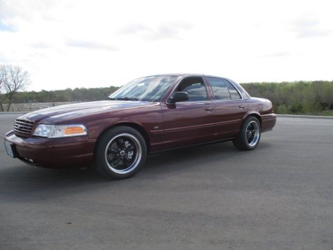 2004 Ford Crown Victoria - A maroon 2004 Ford Crown Victoria is parked on an asphalt surface with a treeline and cloudy sky in the background. The sedan features blac…