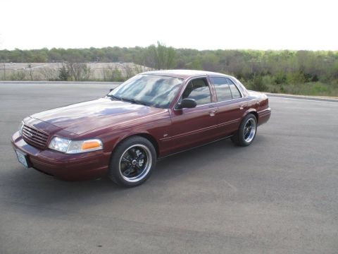 2004 Ford Crown Victoria - A maroon 2004 Ford Crown Victoria is parked on an asphalt surface with a wooded area in the background. The car is viewed from a 3/4 front…