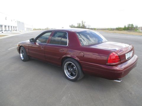 2004 Ford Crown Victoria - The rear passenger side of a dark red 2004 Ford Crown Victoria is shown parked on an asphalt surface. The car has tinted windows and afterm…