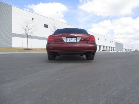 2004 Ford Crown Victoria - The rear view of a dark red Ford Crown Victoria sedan is shown parked in an asphalt lot in front of a large industrial building with multip…