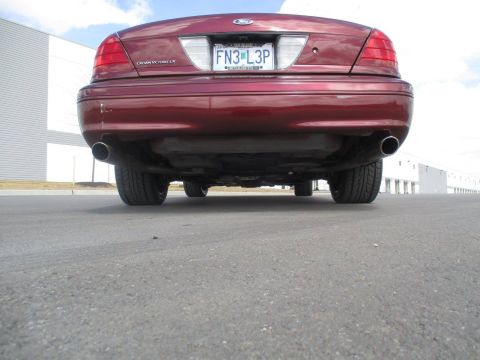 2004 Ford Crown Victoria - Rear view of a dark red sedan parked on asphalt with a warehouse in the background. The vehicle has dual exhaust pipes visible underneath.