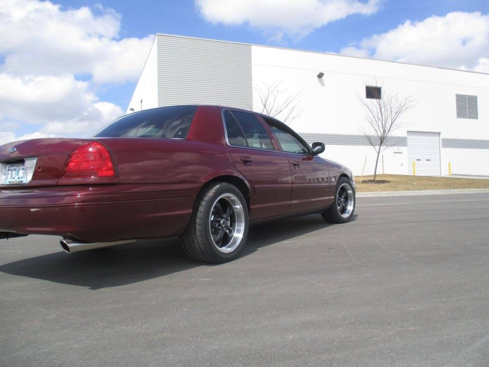 Maroon Ford Crown Victoria sedan, rear passenger side view, with aftermarket wheels parked in front of a building.