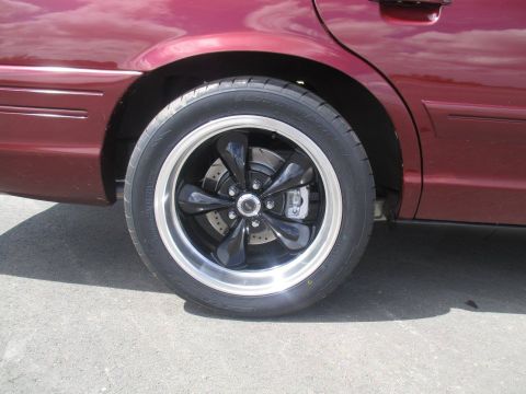 2004 Ford Crown Victoria - Close-up side view of a maroon sedan's rear wheel and fender. The vehicle features a custom black five-spoke wheel with a polished lip, and…
