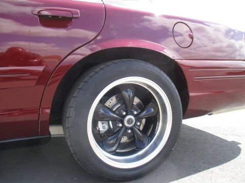 2004 Ford Crown Victoria - The rear passenger side of a dark red sedan is shown, focusing on the wheel and fender area. The vehicle features a shiny black five-spoke…