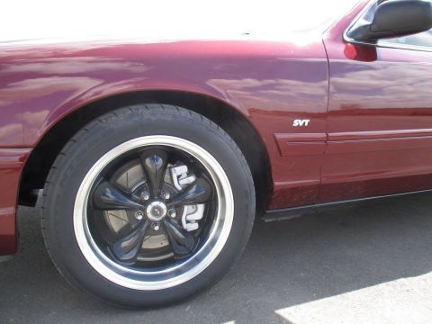 2004 Ford Crown Victoria - A close-up, side-angle view of the front fender and wheel of a dark red sedan. The vehicle features a black five-spoke wheel with a polishe…