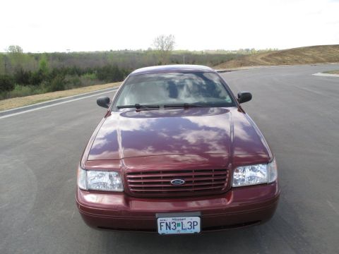 2004 Ford Crown Victoria - Front view of a dark red Ford Crown Victoria parked on an asphalt road with a wooded area and a hill in the background. The car has a chrom…