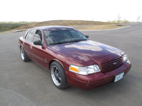 2004 Ford Crown Victoria - A maroon Ford Crown Victoria is parked on an asphalt road with rolling hills and scattered trees in the background. The car is viewed from…