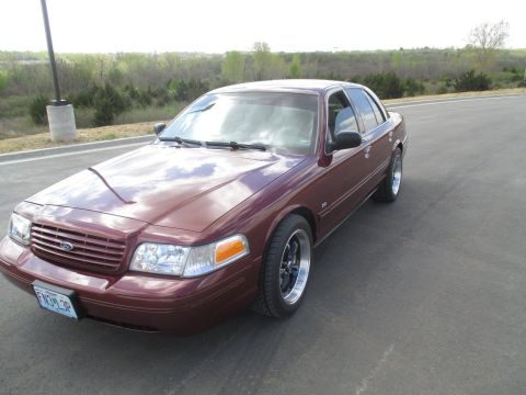2004 Ford Crown Victoria - A maroon 2004 Ford Crown Victoria is parked on an asphalt road with a wooded landscape in the background. The car is viewed from a 3/4 fron…