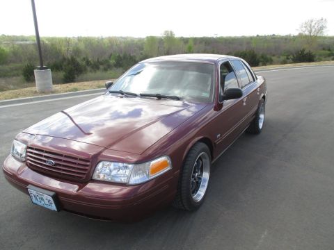 2004 Ford Crown Victoria - A maroon 2004 Ford Crown Victoria is parked on an asphalt road with a tree line and sky in the background. The car is viewed from a three-q…