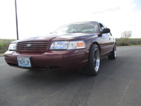 2004 Ford Crown Victoria - A low-angle, front-three-quarter view of a maroon Ford Crown Victoria sedan parked on an asphalt surface. The car features a chrome grille,…