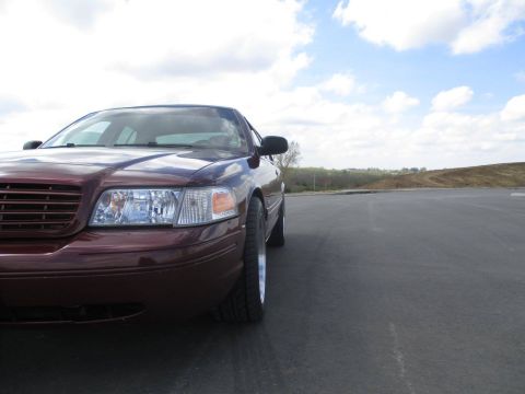 2004 Ford Crown Victoria - The front passenger side of a maroon Ford Crown Victoria is shown parked on an asphalt lot with a blue sky and clouds overhead.