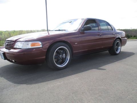2004 Ford Crown Victoria - A maroon sedan with aftermarket black and chrome wheels is parked on an asphalt surface with a blurred background of trees and sky. The veh…