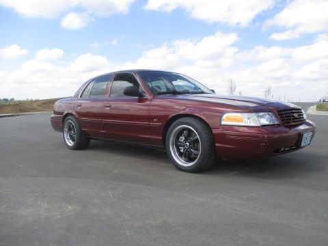 2004 Ford Crown Victoria - A maroon 2004 Ford Crown Victoria sedan is parked on an asphalt surface with a cloudy blue sky in the background. The car features aftermar…