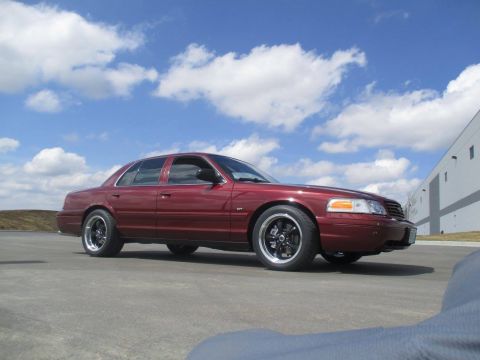 2004 Ford Crown Victoria - A maroon 2004 Ford Crown Victoria is parked on an asphalt lot with a building and cloudy sky in the background. The sedan features custom b…
