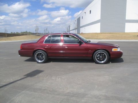 2004 Ford Crown Victoria - Side view of a maroon Ford Crown Victoria sedan parked on asphalt in front of a modern industrial building. The car has aftermarket black w…