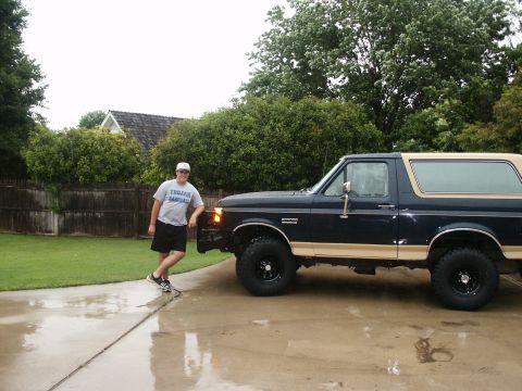 1991 Ford Bronco - A dark navy blue two-tone Ford Bronco full-size SUV with tan lower panels sits in profile on a wet driveway, lifted on black wheels with la…