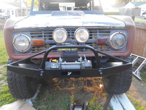 1972 Ford Bronco - Front view of a weathered, reddish-brown Ford Bronco with a black aftermarket bumper and winch. The vehicle has two large round headlights…