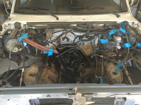1987 Ford Bronco - The engine bay of a white Ford Bronco is shown with the engine partially disassembled. Various hoses, wires, and components are visible, ma…