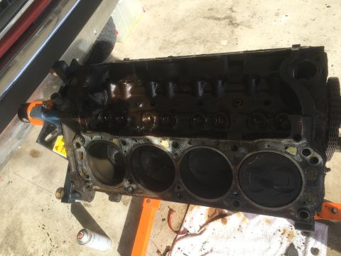 1987 Ford Bronco - A heavily soiled V8 engine block is shown mounted on an orange engine stand. The cylinder bores are visible, with pistons in some of them,…