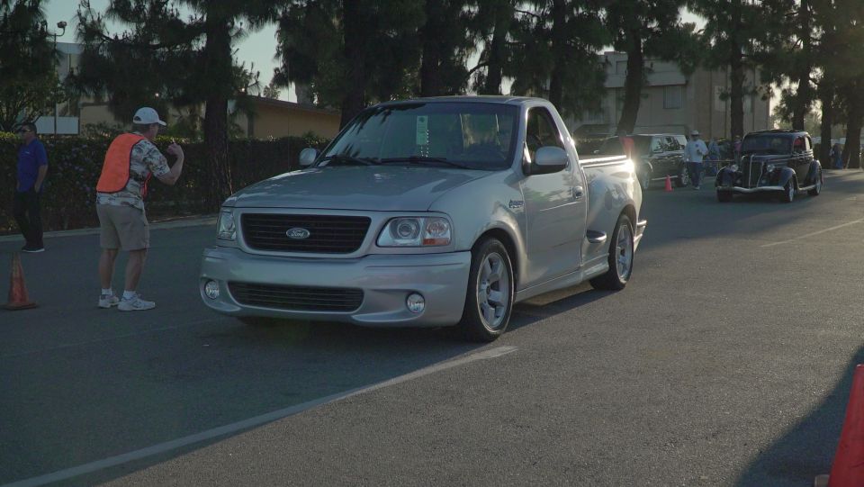 Silver Ford F-150 Lightning pickup truck parked on asphalt with a vintage black Ford coupe nearby.
