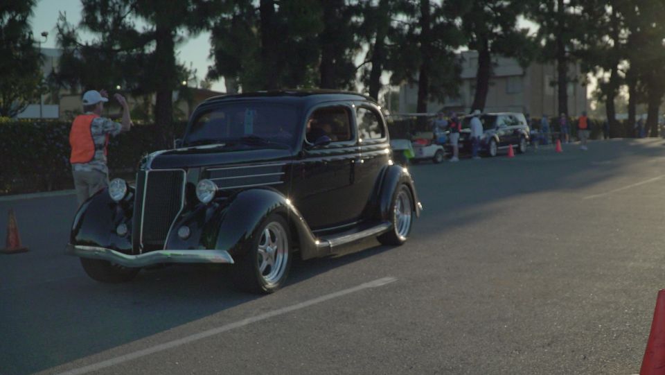 Black classic hot rod sedan parked on asphalt road with people and other vehicles in background.