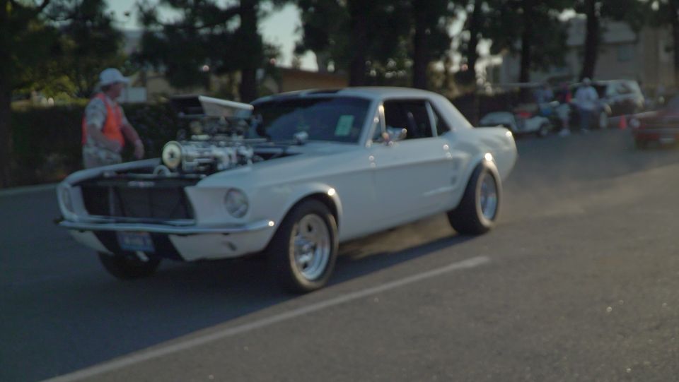 White 1960s Ford Mustang with exposed engine and a person in an orange vest.