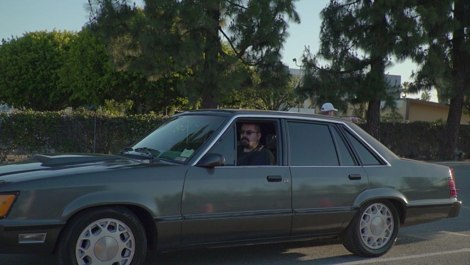 Man driving a dark green sedan, with another person leaning out of the rear window.